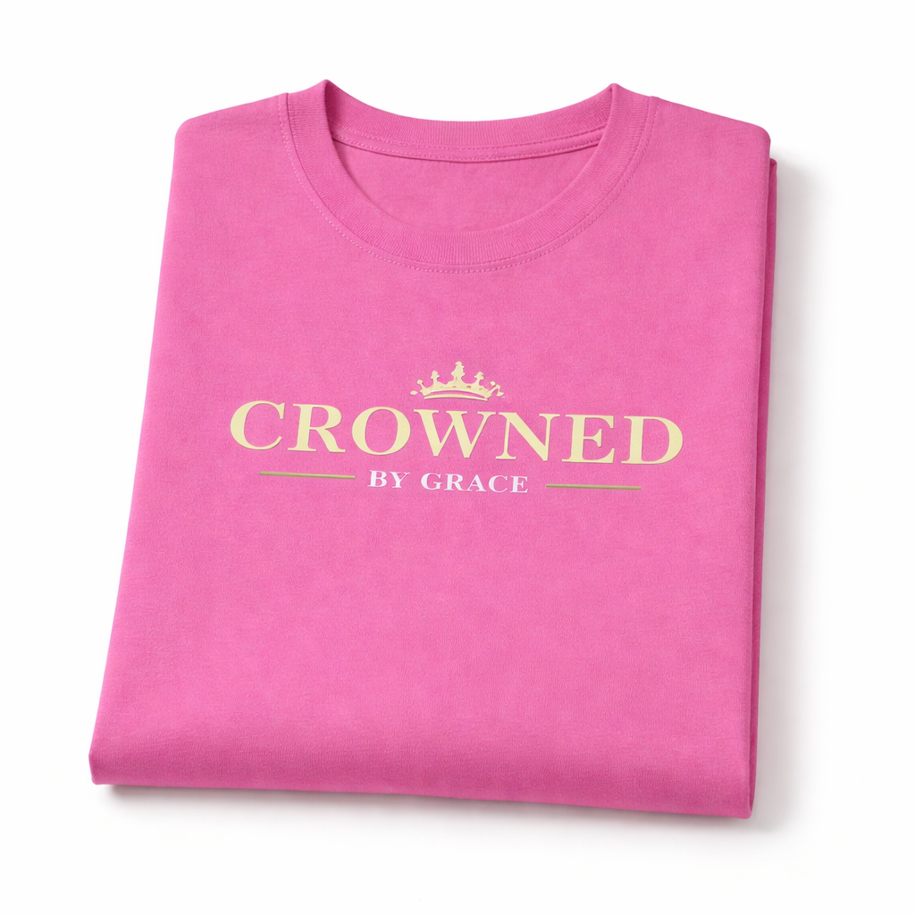 Folded pink t-shirt with 'Crowned by Grace' text on a white background