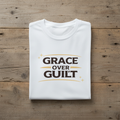 Folded white t-shirt with 'Grace Over Guilt' text on a wooden surface