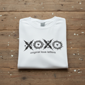 Folded Original Love Letters Christian Believers white t-shirt with black graphic design and text on a wooden surface