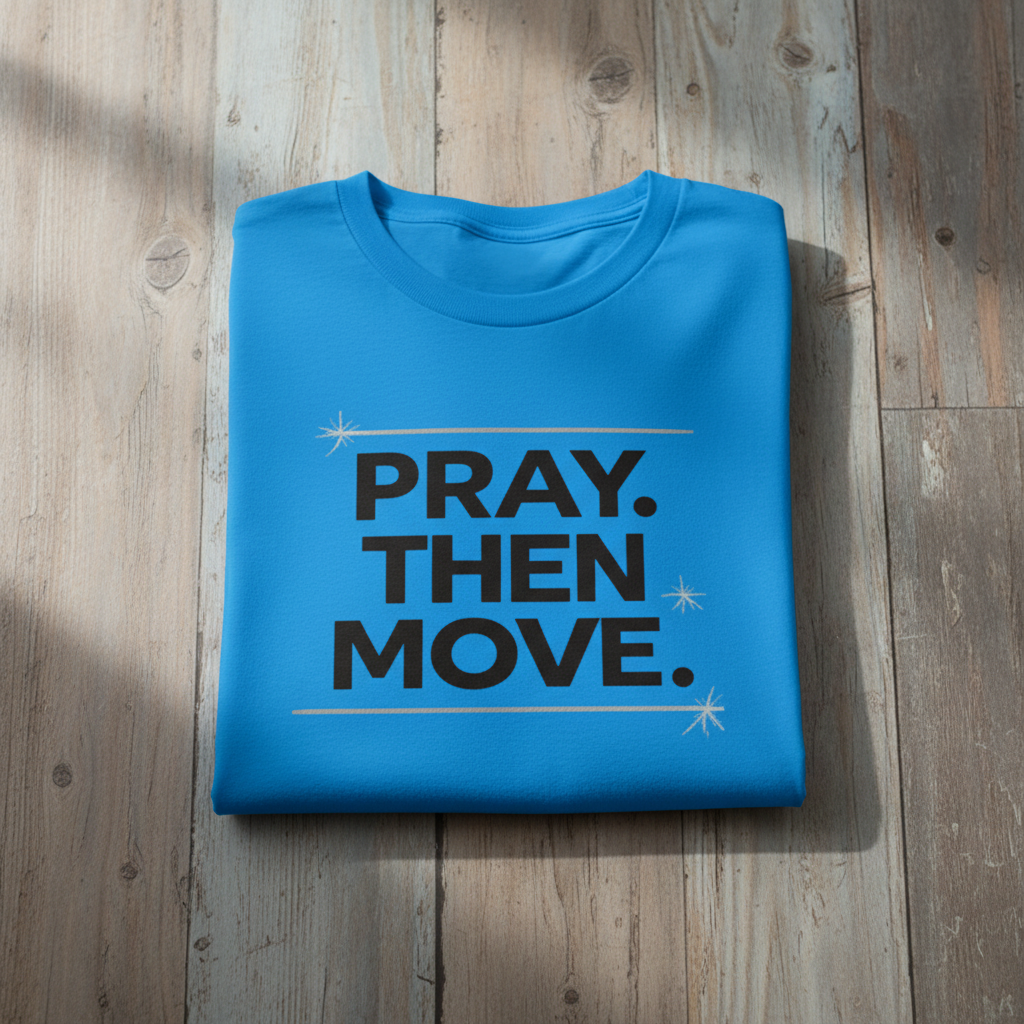 Folded blue t-shirt with 'PRAY. THEN MOVE.' text on a wooden surface